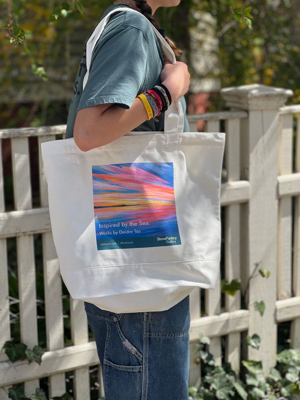 A picture of a woman carrying a large canvas tote on her shoulder. The tote bag has a colorful painting of a sunset with pink, blue, and orange brushstrokes and the words "Inspired by the Sea: Works by Deidre Tao, StoveFactory Gallery" and the artist's website and social media handle in small lettering at the base of the bag.